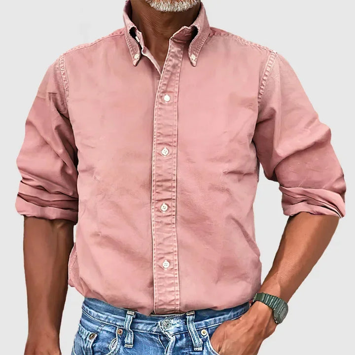 Man wearing a pink button-up shirt with rolled sleeves and blue jeans against a plain gray background.