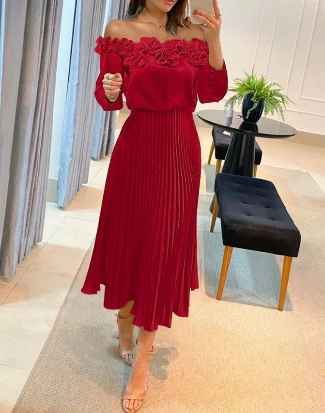 Woman in red off-shoulder maxi dress with ruffled neckline and pleated skirt, standing in modern indoor setting with chairs and plant.