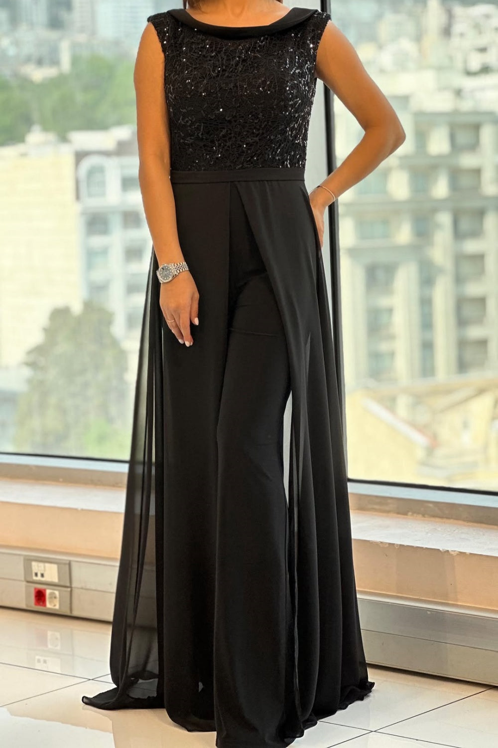 Woman in black textured tulle jumpsuit with flowing legs, standing indoors near a large window with cityscape view.
