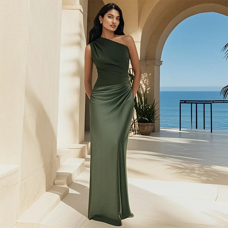 Woman in a green one-shoulder maxi dress standing in a sunlit architectural balcony overlooking the sea.