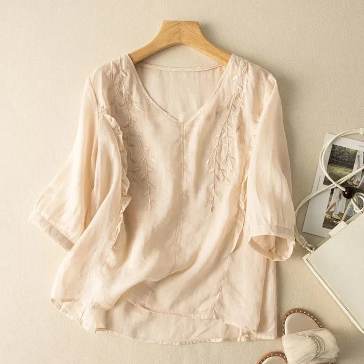 Cream loose retro top with delicate leaf embroidery, on a wooden hanger against a neutral wall, with sandals and bag.
