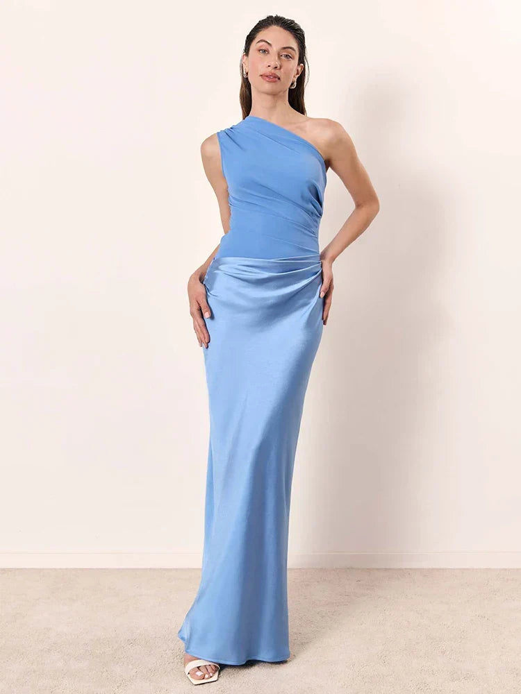 Woman in a light blue one-shoulder maxi dress posing against a plain neutral indoor background.