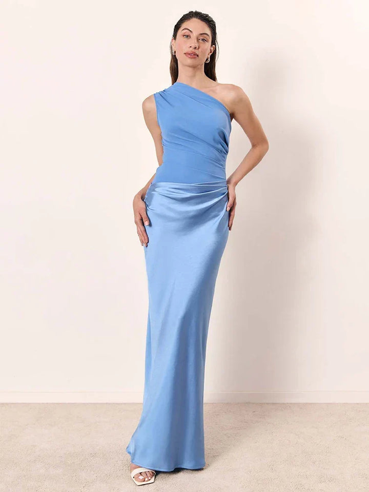 Woman in a light blue one-shoulder maxi dress posing against a plain neutral indoor background.