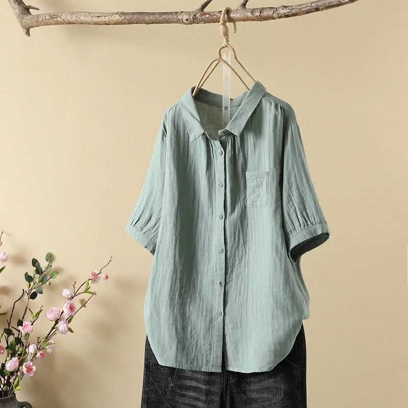 Mint green casual loose blouse hanging on a wooden branch, with pink flowers nearby, against a light beige background.