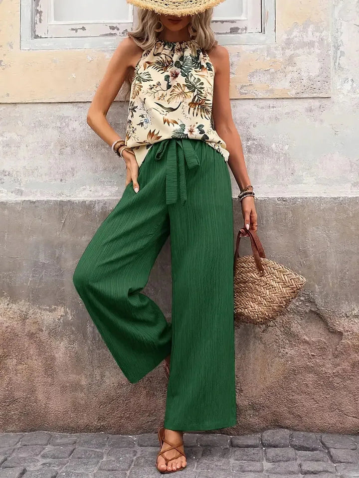 Woman in a floral sleeveless top and green wide-leg pants, accessorized with a straw hat and woven bag, in front of a textured wall.