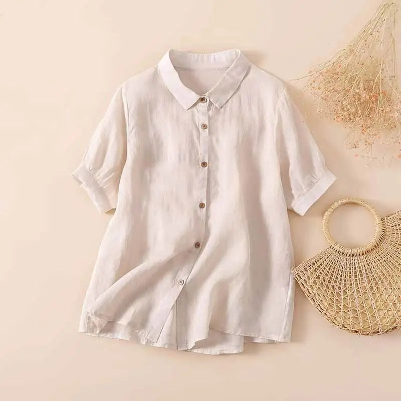 White casual button-up shirt with short sleeves, displayed on a neutral background next to a woven handbag and dried flowers.