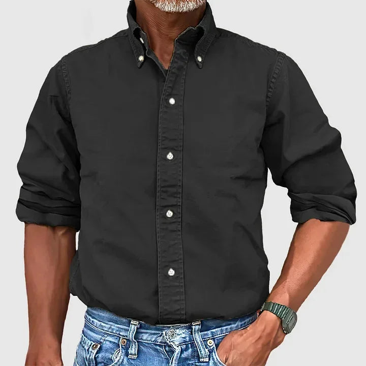 Man wearing a black button-up shirt with rolled sleeves and blue jeans against a plain gray background.