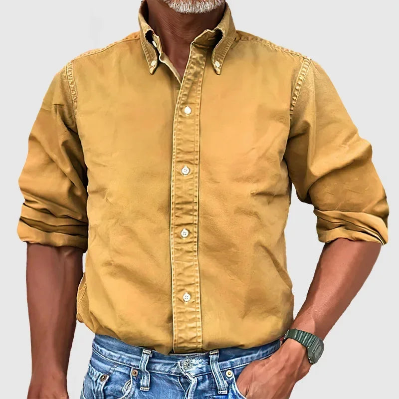 Man wearing a mustard yellow button-up shirt with rolled sleeves and blue jeans against a plain gray background.
