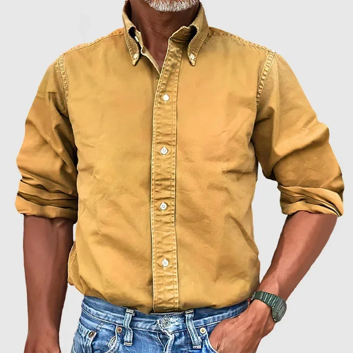 Man wearing a mustard yellow button-up shirt with rolled sleeves and blue jeans against a plain gray background.
