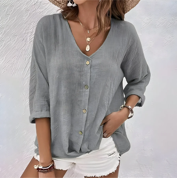 Woman in a gray relaxed button-up blouse with rolled sleeves, accessorized with jewelry, set against a textured white background.