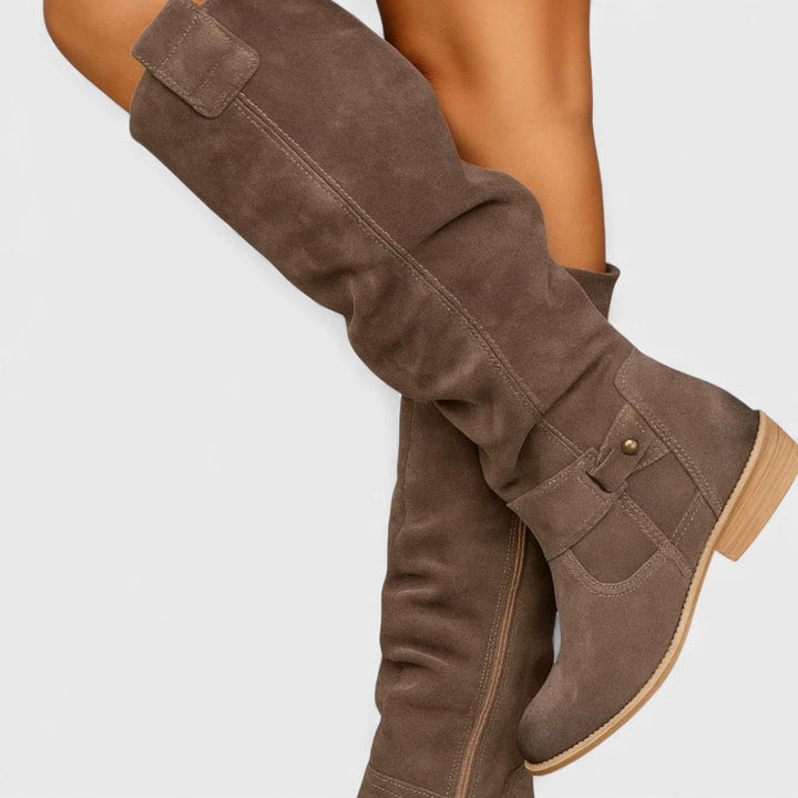 Person wearing brown knee-high boots with buckle detail and wooden heels, studio background.