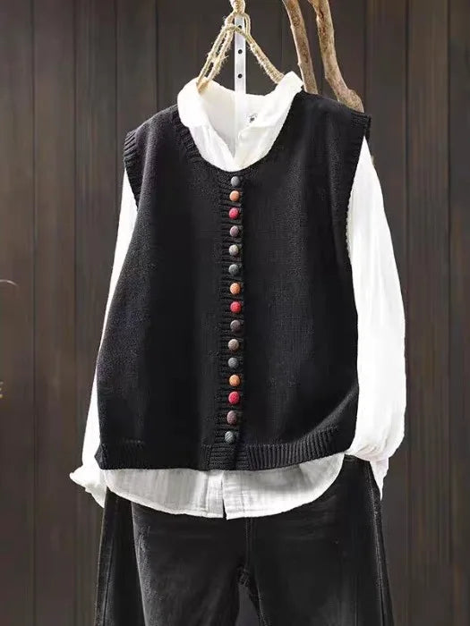 Black sleeveless cardigan with colorful buttons over white blouse hung on dark wooden background.