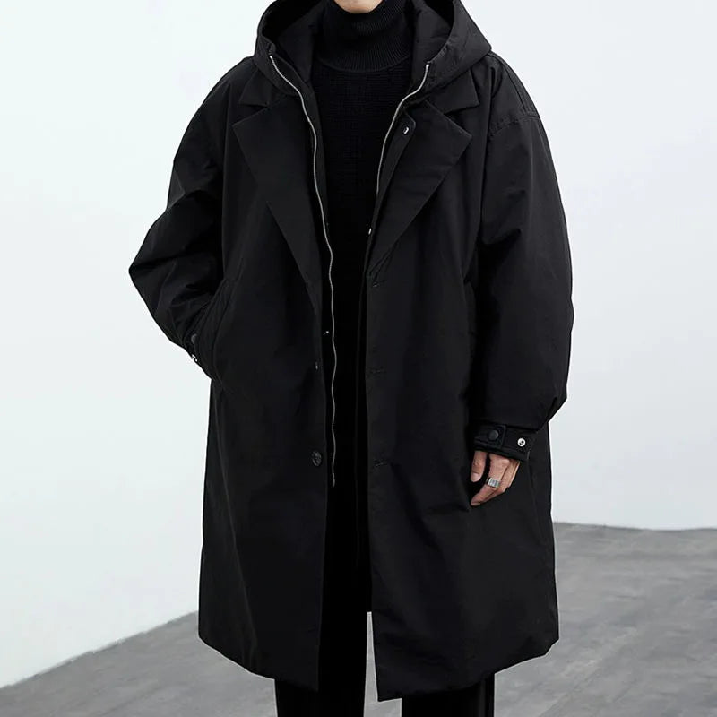 Front view of a black long coat with hood, hands in pockets, in a gray studio.