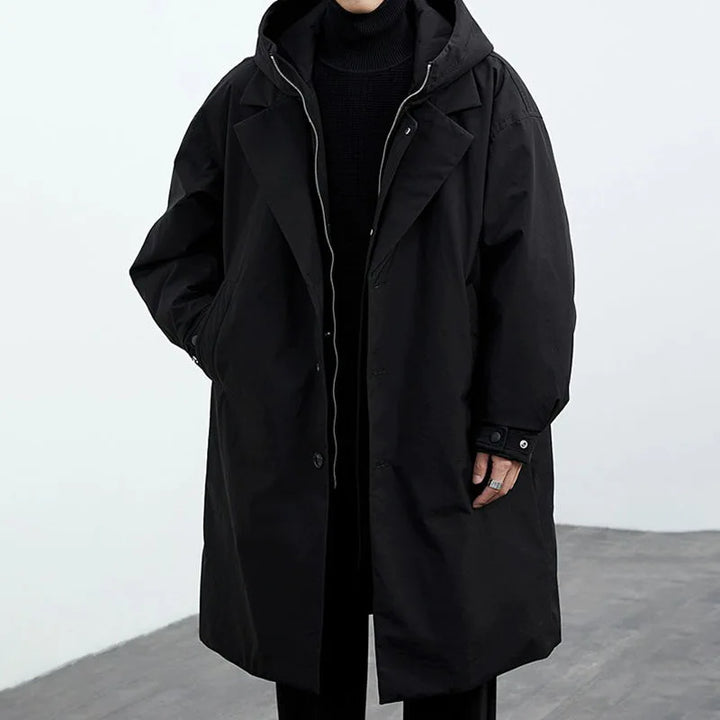 Front view of a black long coat with hood, hands in pockets, in a gray studio.