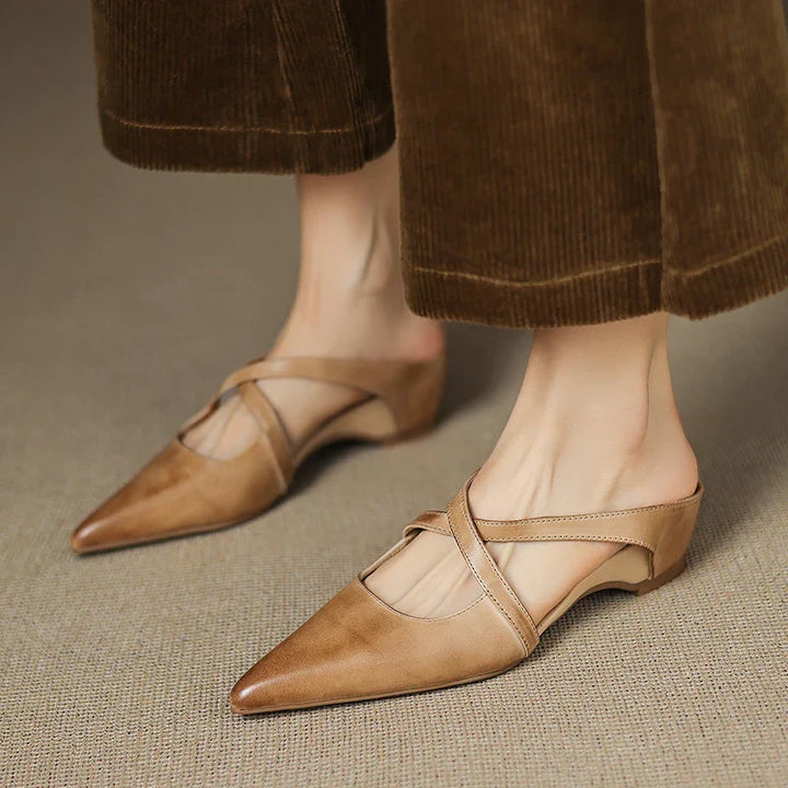 Person wearing tan mules with criss-cross straps on a textured surface.