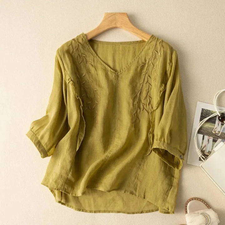 Green loose retro top with subtle leaf embroidery, hung on a wooden hanger against a neutral wall, with sandals and bag nearby.