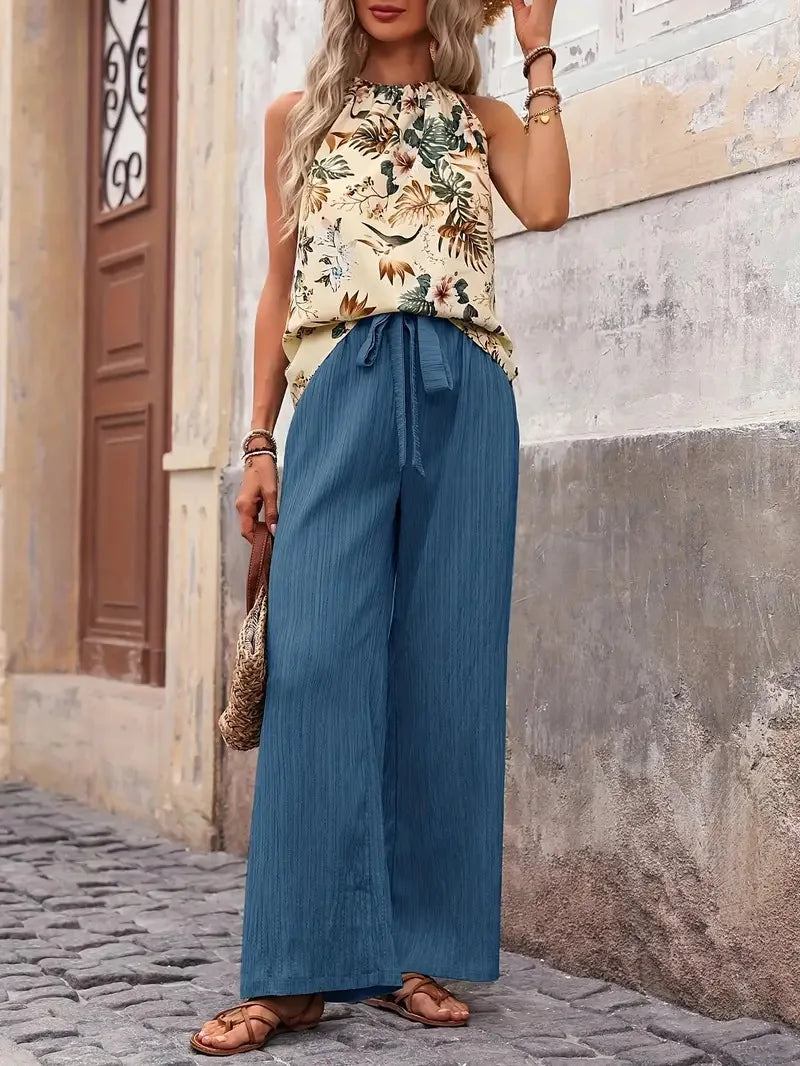 Model in a sleeveless floral top with blue wide-leg pants, standing next to a rustic building wall.