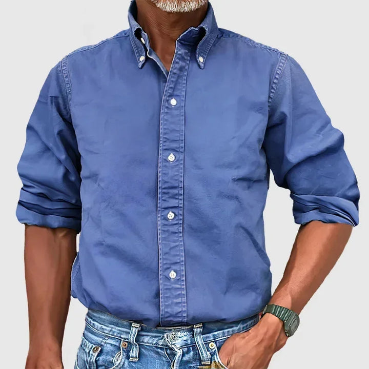 Man wearing a royal blue button-up shirt with rolled sleeves and blue jeans against a plain gray background.