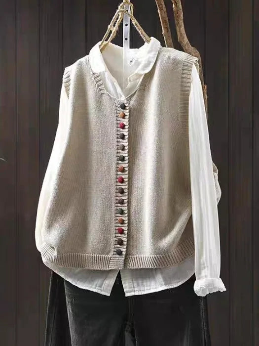 Beige sleeveless cardigan with colorful buttons over white blouse hung on dark wooden background.