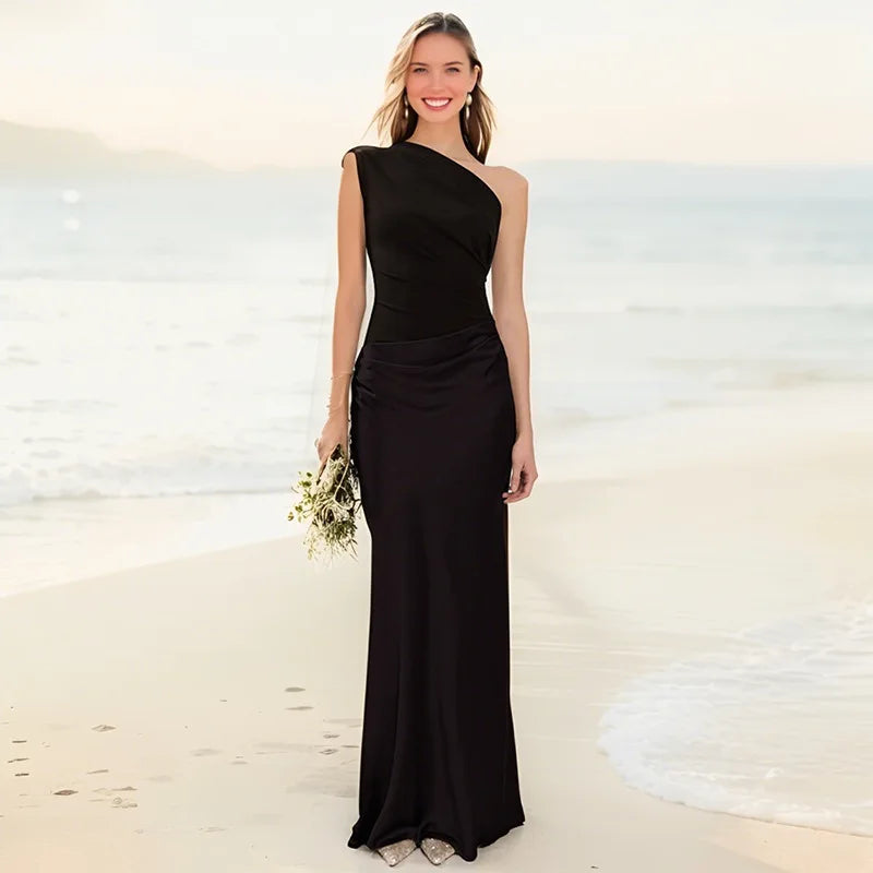 Woman in a black one-shoulder maxi dress holding flowers on a sunny beach with gentle waves in the background.