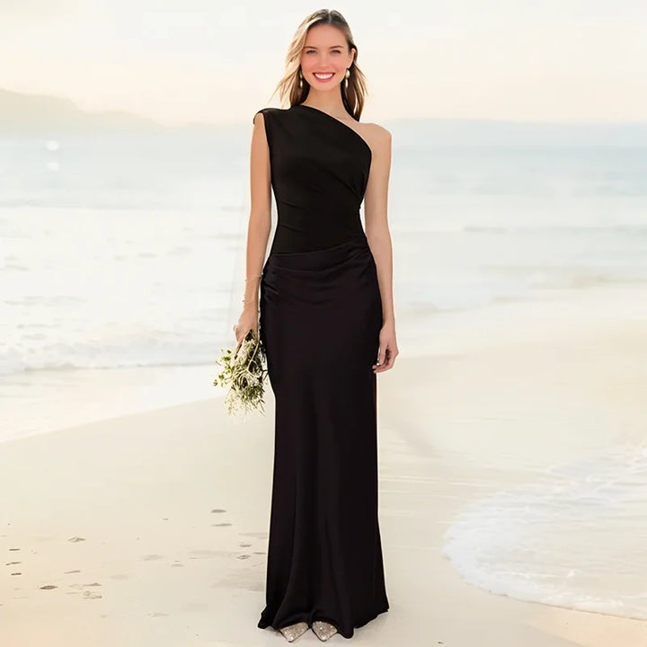 Woman in a black one-shoulder maxi dress holding flowers on a sunny beach with gentle waves in the background.