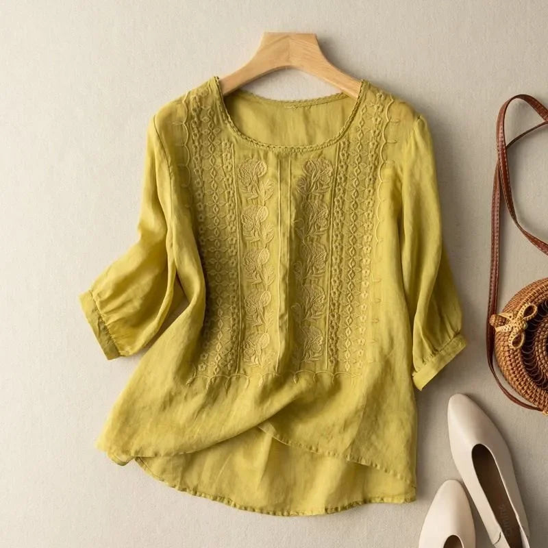 Yellow elegant soft blouse with ornate embroidery on hanger, with shoes and bag against a neutral wall.