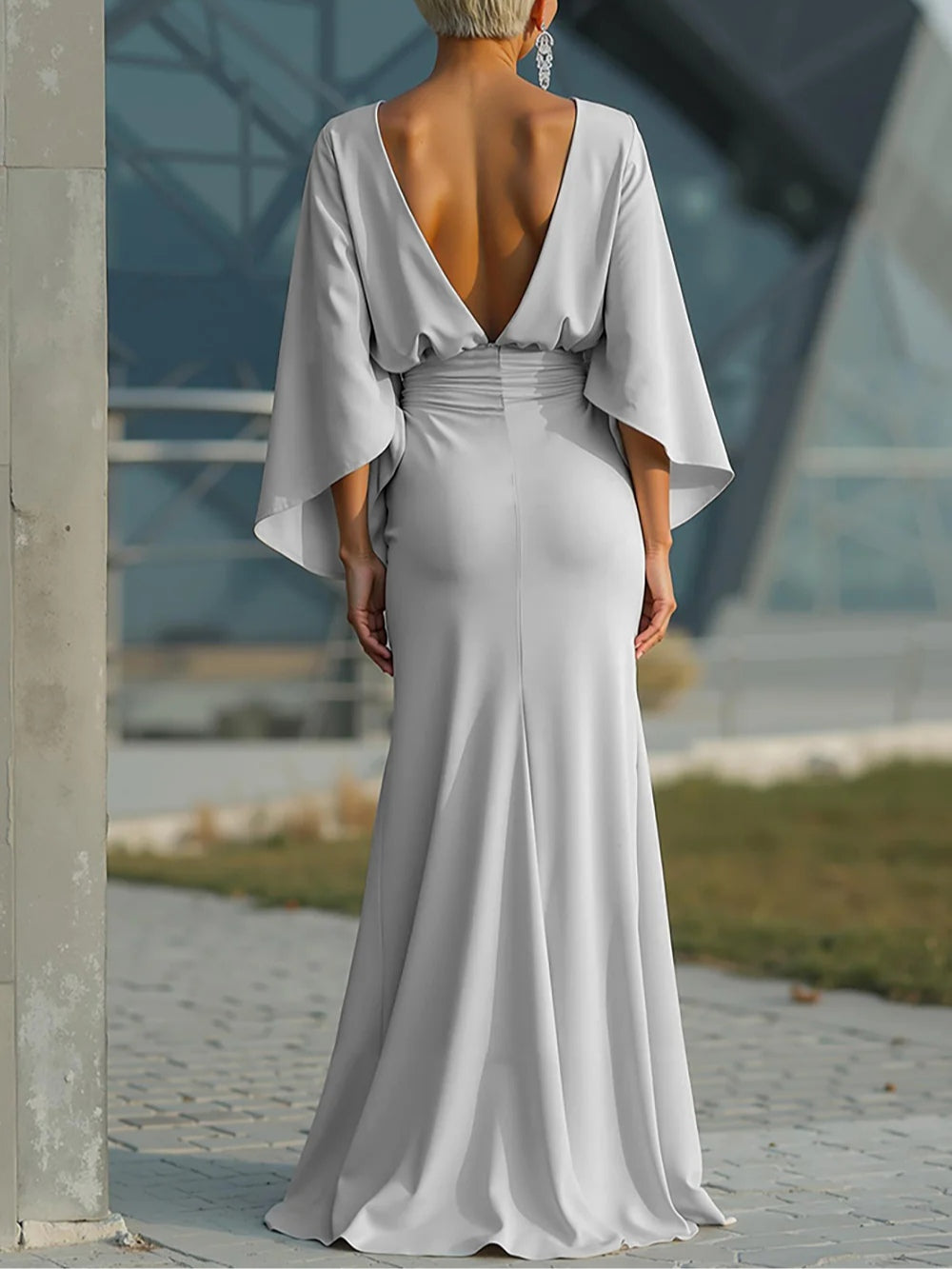 Back view of elegant silver dress showcasing deep V-back and flowing fit, worn by a model outdoors near modern architecture.