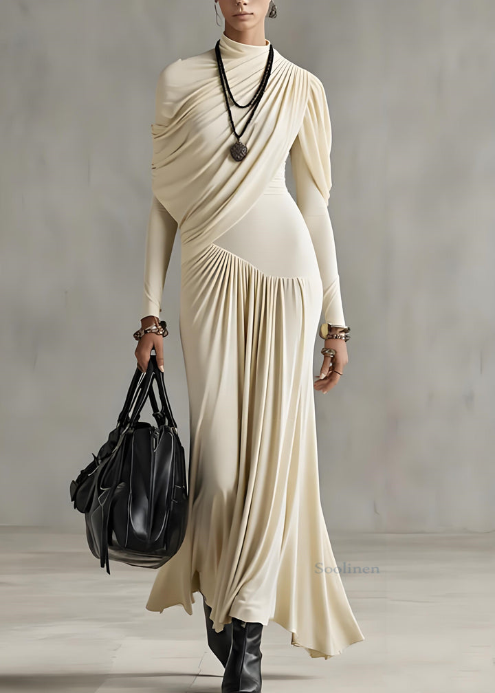 Woman in stylish apricot long dress with draped details, holding a black bag, set in a minimalist indoor background.