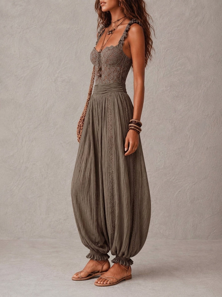 Side view of a woman in a detailed lace top and flowy pleated pants, accessorized with a woven bag and sandals, in a studio setting.