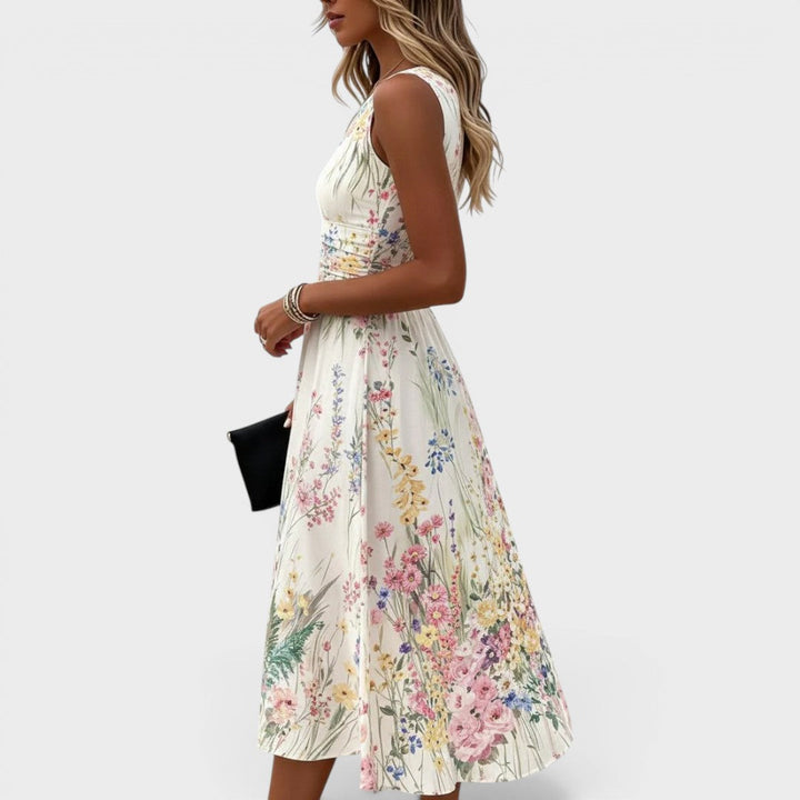 Side view of sleeveless floral midi dress, fitted waist, flowing skirt. Model holding clutch, plain background.