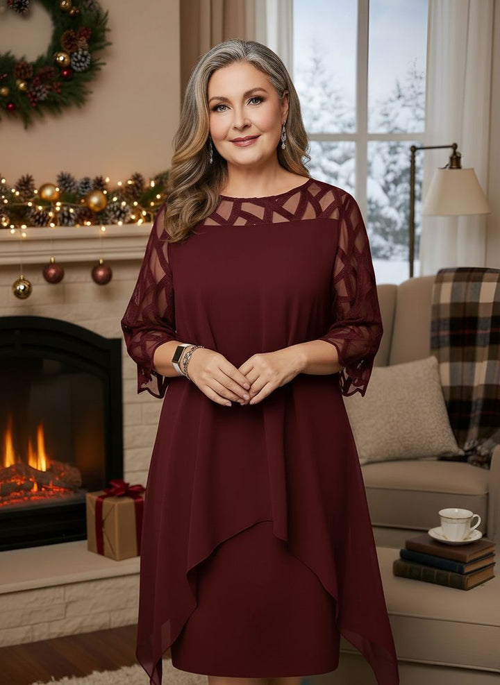 Woman in elegant maroon dress with lace sleeves, standing indoors by decorated fireplace.