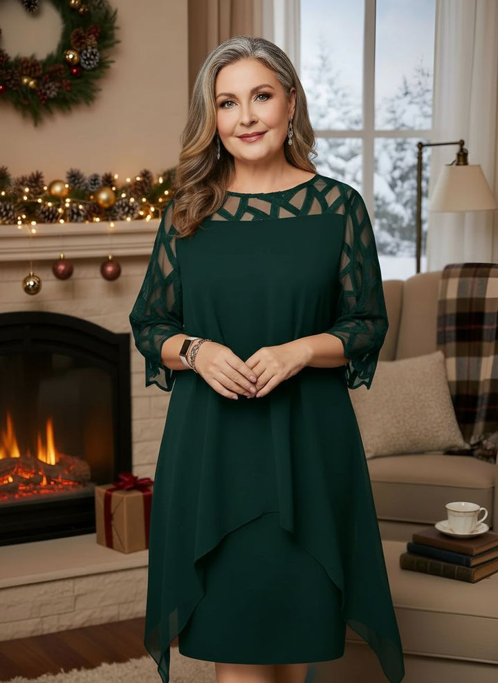 Woman in elegant emerald green dress with lace sleeves, standing indoors by decorated fireplace.