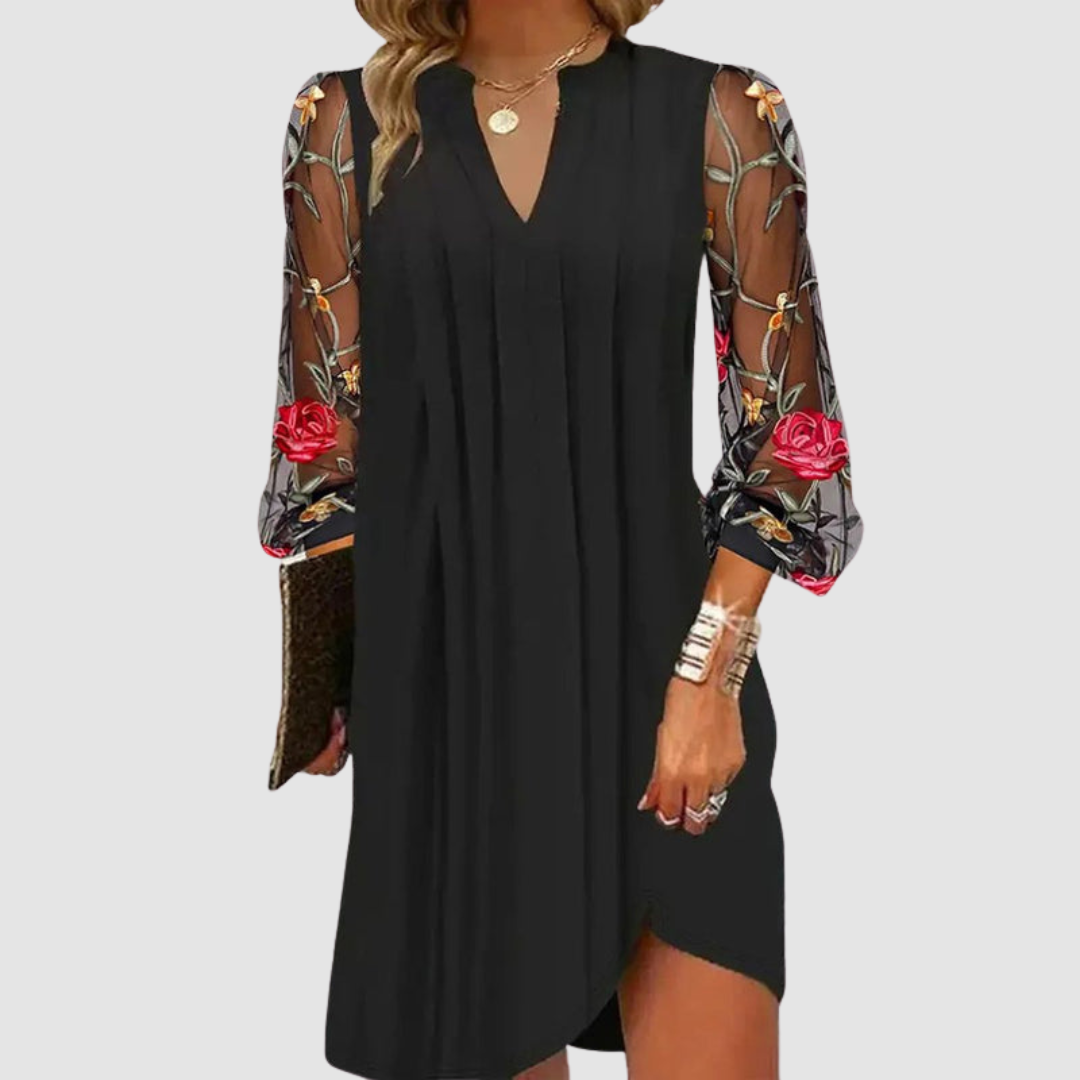 Close-up of a black dress with sheer floral embroidered sleeves, worn by a model holding a clutch, on a white background.