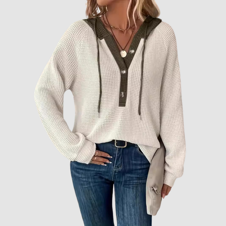 White knitted long sleeve hoodie with button details and drawstrings, worn with blue jeans against a gray studio background.