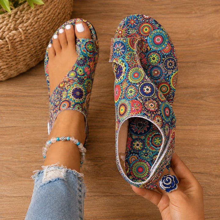 Circular-patterned colorful orthopedic sandals on feet with anklet, denim jeans, wooden floor background.