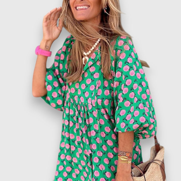 Close-up of woman in green dress with pink floral pattern, smiling and touching her hair, on plain background.