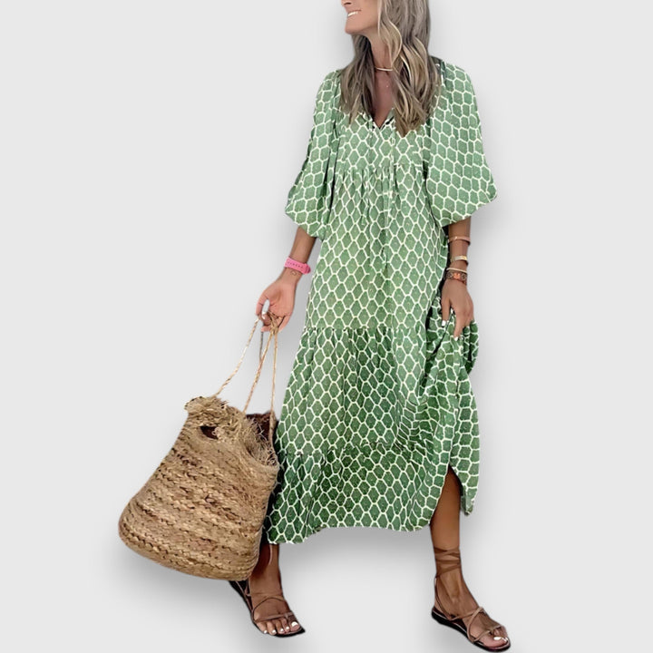Woman in green geometric-patterned dress, standing with a woven tote, on plain background.