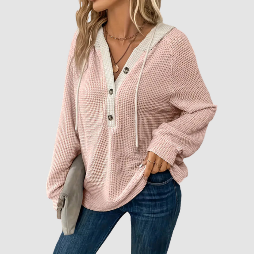 Pink knitted long sleeve hoodie with button details and drawstrings, paired with blue jeans in a gray studio setting.
