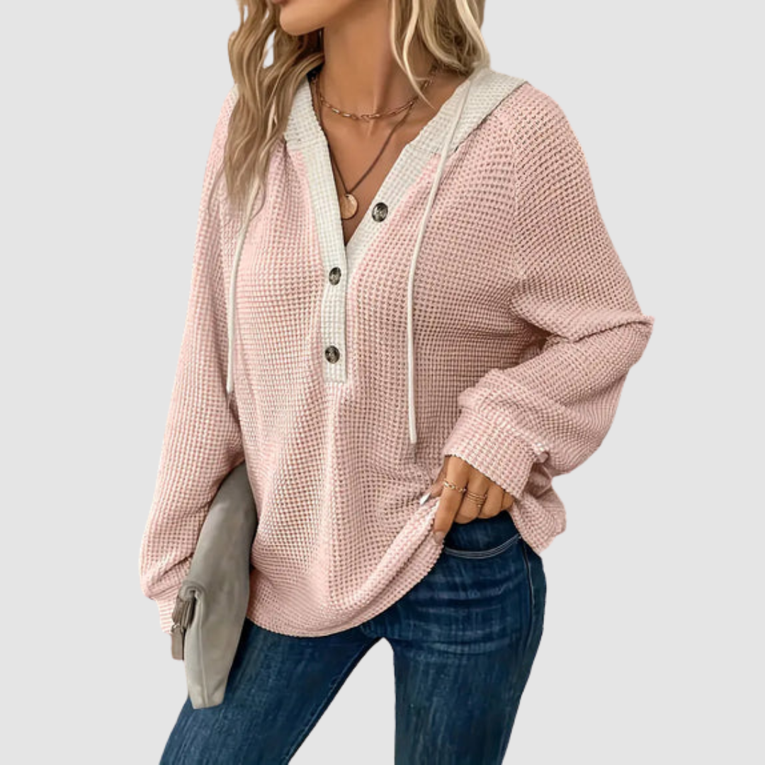 Pink knitted long sleeve hoodie with button details and drawstrings, paired with blue jeans in a gray studio setting.