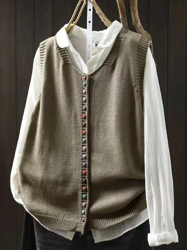 Olive sleeveless cardigan with colorful buttons over white blouse hung on dark wooden background.