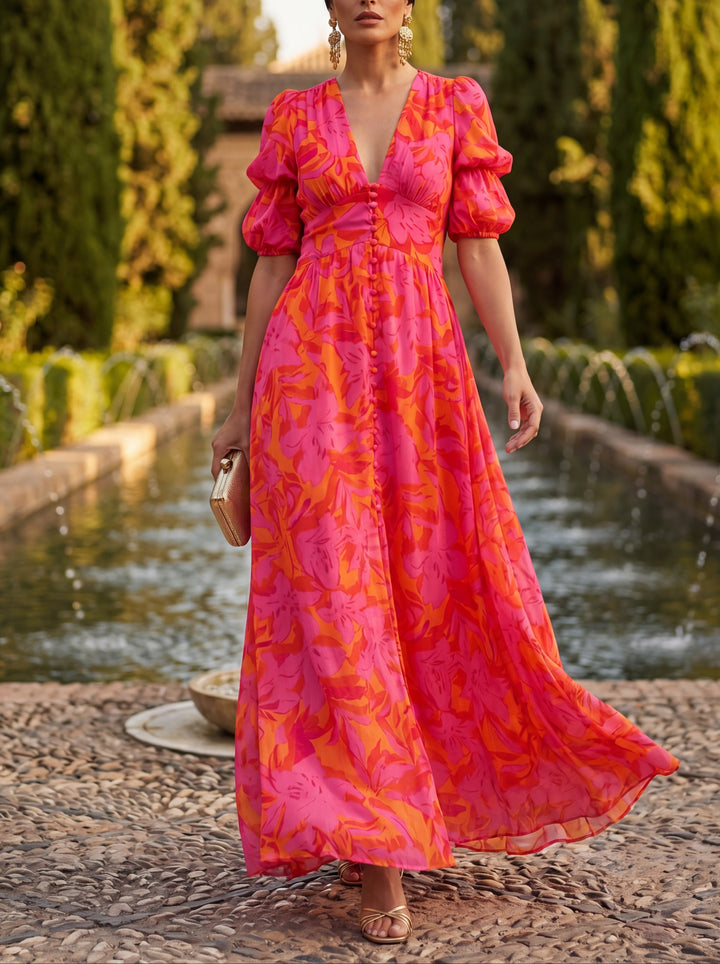 Woman in vibrant pink and orange flower dress walking outdoors by a fountain, surrounded by lush greenery.