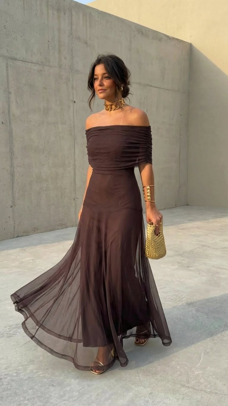 Woman wearing a brown off-shoulder dress with a sheer flowing skirt, holding a gold clutch, standing on a concrete outdoor surface.