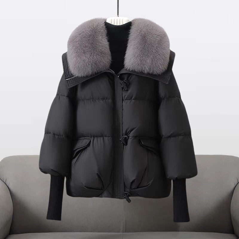 Black winter jacket with a large fluffy gray collar displayed on a sofa in a studio setting.