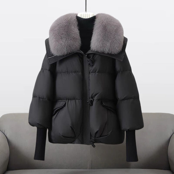 Black winter jacket with a large fluffy gray collar displayed on a sofa in a studio setting.
