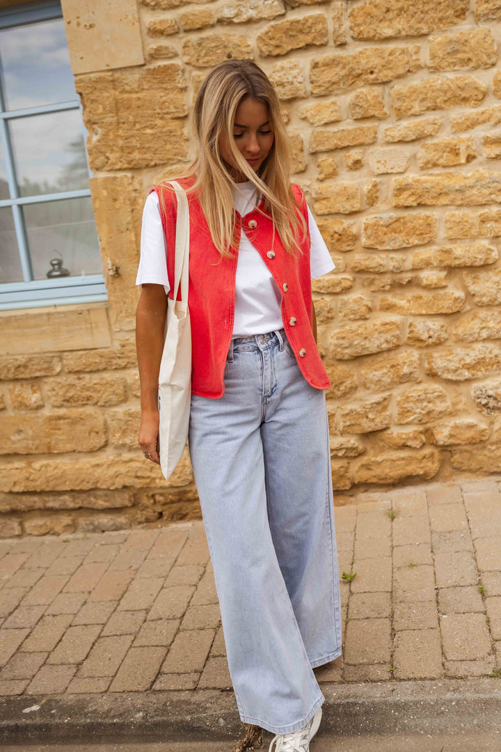 Woman in red vest, white shirt, light jeans, and sneakers, posing outdoors with stone wall and window as background.