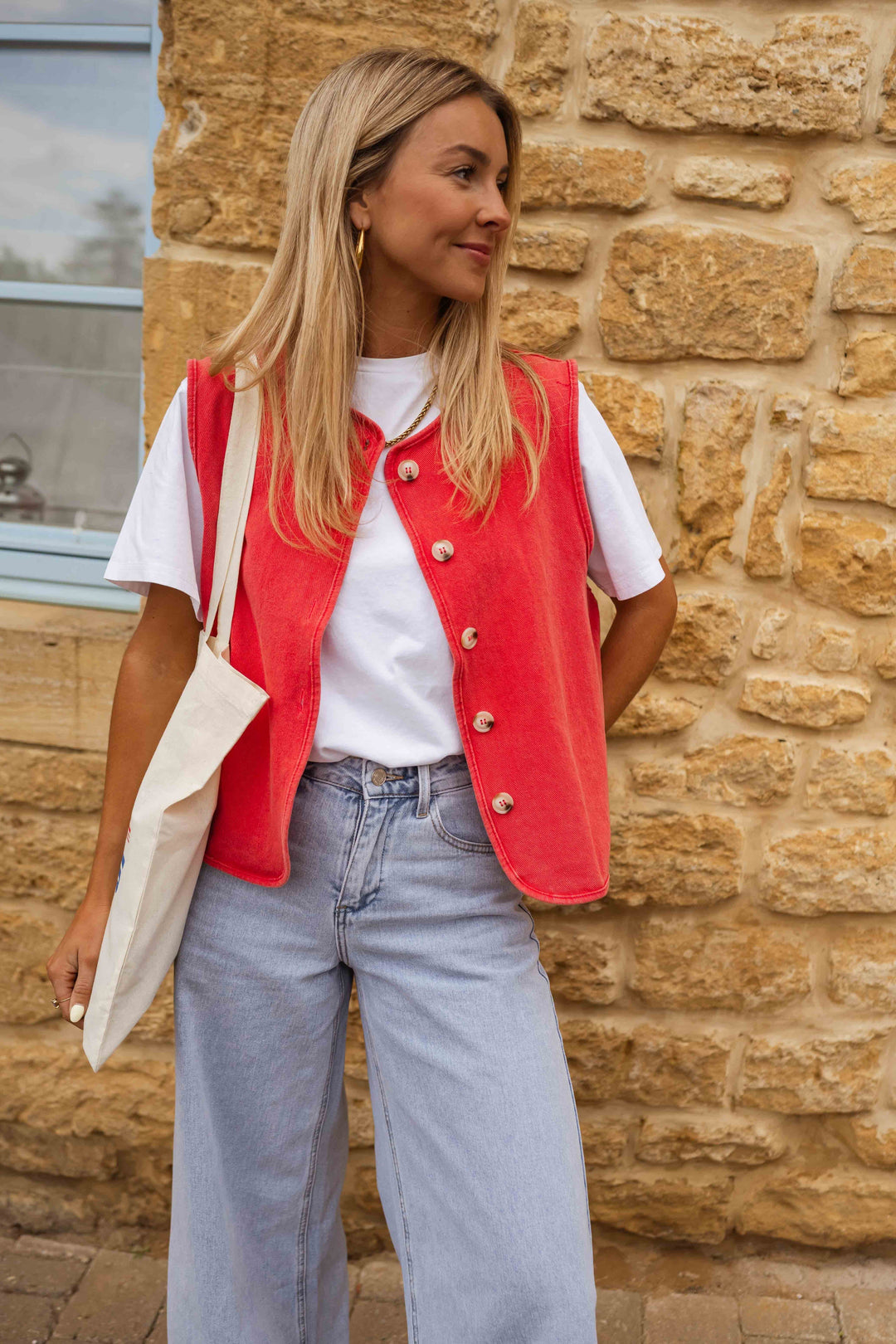 Woman wearing red buttoned vest over white shirt and light jeans, standing against stone wall with a tote bag.