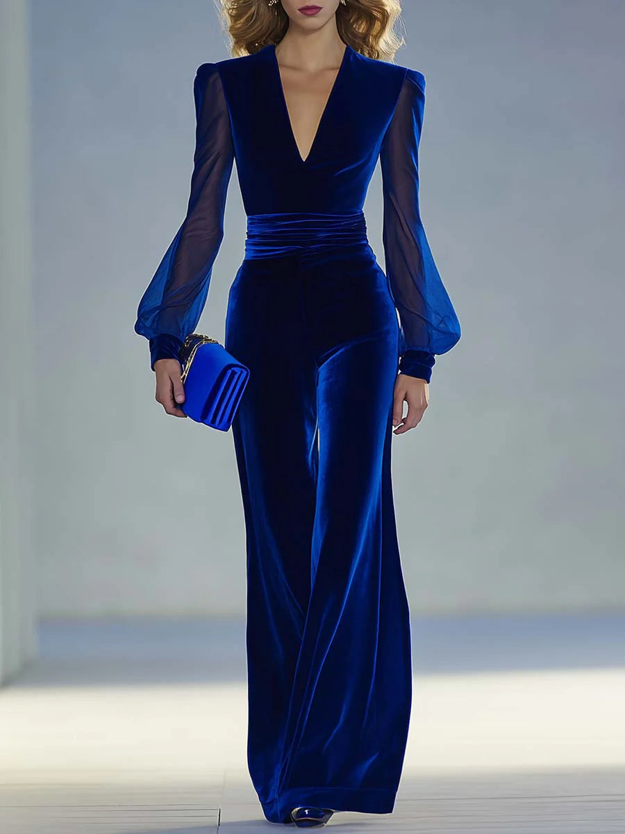 Elegant blue suede V-neck jumpsuit with sheer sleeves worn by a model holding a clutch in a softly lit studio.