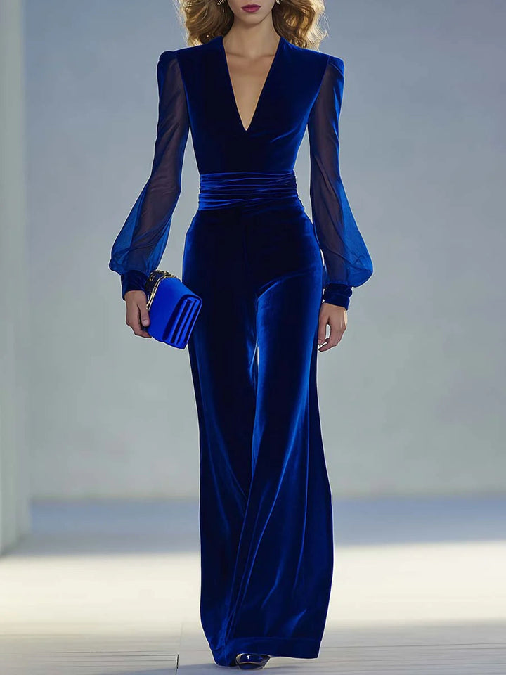 Elegant blue suede V-neck jumpsuit with sheer sleeves worn by a model holding a clutch in a softly lit studio.