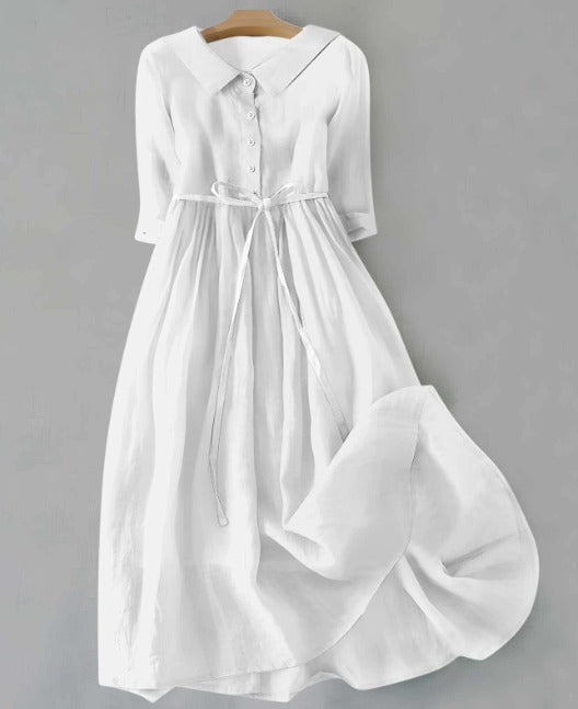 White short sleeve dress with buttons and tie waist, hanging on a wooden hanger against a gray background.