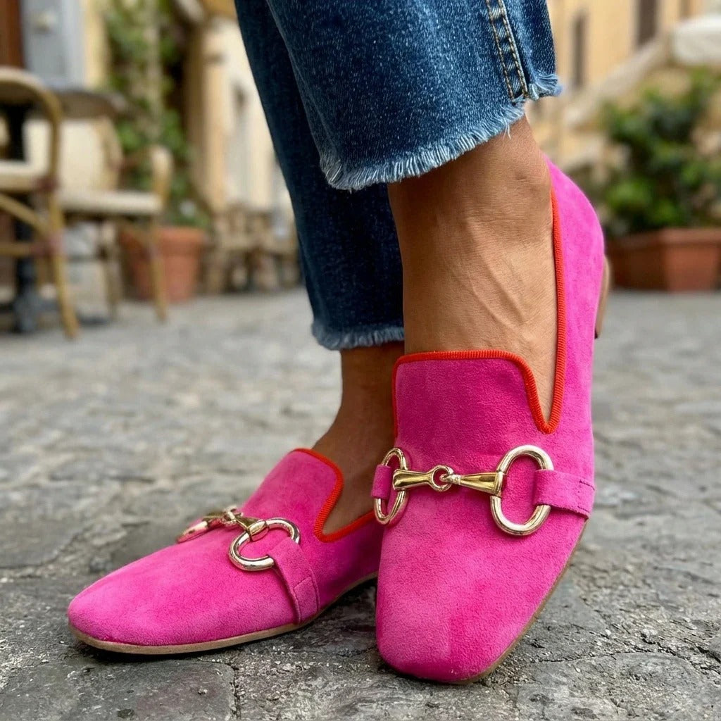 Bright pink loafers with metal detail, showcased against outdoor urban backdrop with plants.